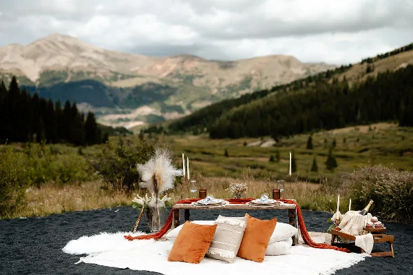 A luxury outdoor picnic setup featuring a wooden table, candles, and pillows on a rug, set against a mountain landscape.