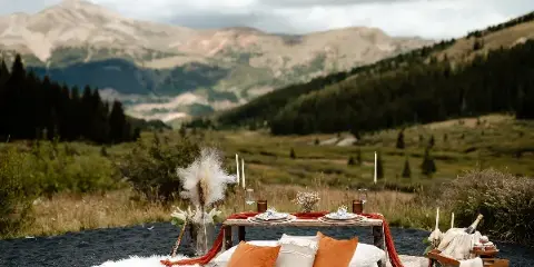 A luxury outdoor picnic setup featuring a wooden table, candles, and pillows on a rug, set against a mountain landscape.