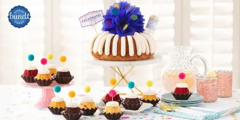 A decorated Bundt cake with white icing and a blue paper flower sits on a stand, surrounded by assorted Bundtinis and party supplies.