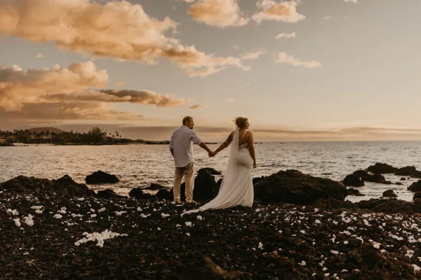A bride and groom holding hands on a rocky beach at sunset.