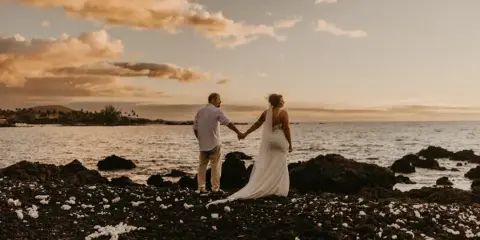 A bride and groom holding hands on a rocky beach at sunset.