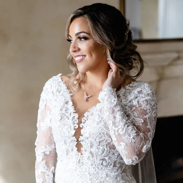 A smiling bride with styled hair and makeup wearing a white lace wedding dress.