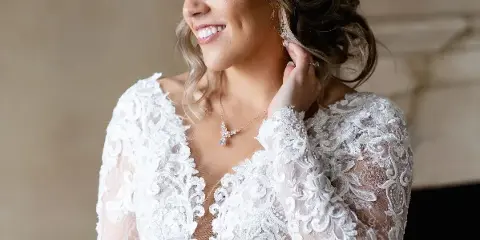 A smiling bride with styled hair and makeup wearing a white lace wedding dress.