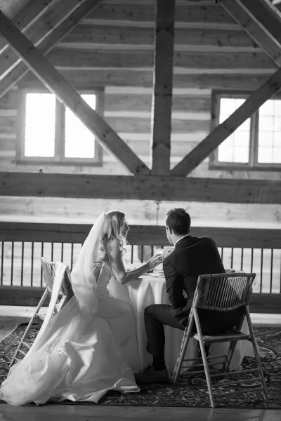 A black and white photograph of a bride and groom sitting at a table in a rustic wooden venue.