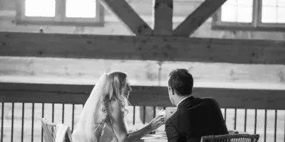 A black and white photograph of a bride and groom sitting at a table in a rustic wooden venue.