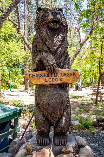 A wooden bear sculpture holding a sign for Christopher Creek Lodge in a forested area.