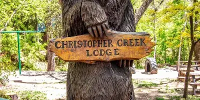 A wooden bear sculpture holding a sign for Christopher Creek Lodge in a forested area.