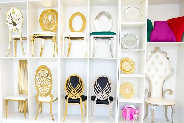 A white shelving unit displaying an assortment of gold and silver chairs, decorative plates, and colorful velvet stools.