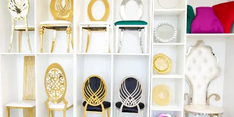 A white shelving unit displaying an assortment of gold and silver chairs, decorative plates, and colorful velvet stools.