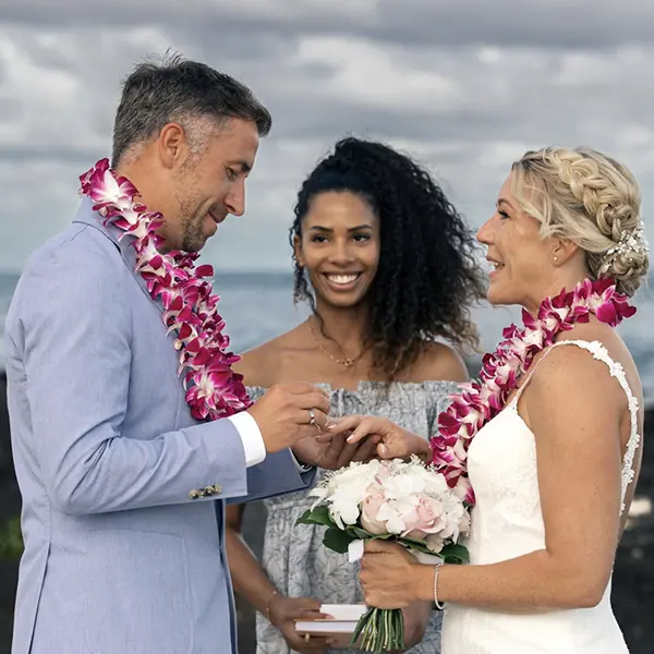 A couple exchanging rings during a beach wedding ceremony, wearing pink and white flower leis, with an officiant standing between them.