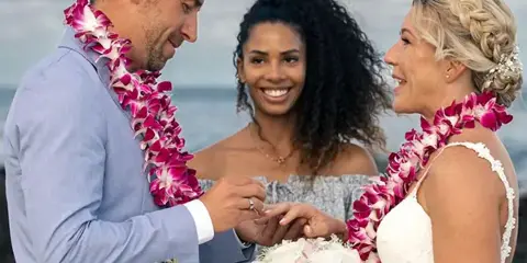 A couple exchanging rings during a beach wedding ceremony, wearing pink and white flower leis, with an officiant standing between them.