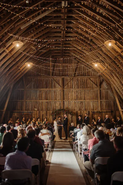 Interior of a wooden barn venue with string lights and a wedding ceremony in progress.
