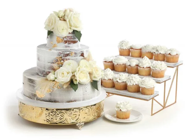 A three-tiered white wedding cake decorated with roses and gold leaf, displayed next to a tiered stand of white frosted cupcakes.