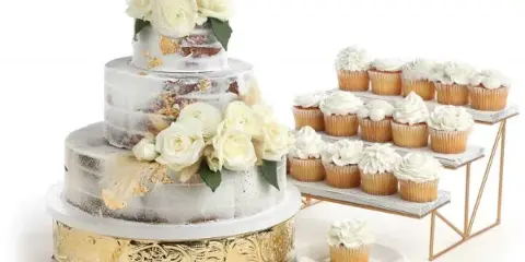 A three-tiered white wedding cake decorated with roses and gold leaf, displayed next to a tiered stand of white frosted cupcakes.