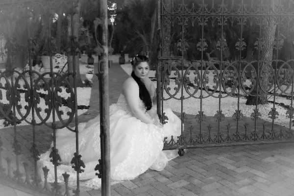 A black and white photograph of a bride in a white wedding dress sitting on a brick path in front of decorative iron gates.