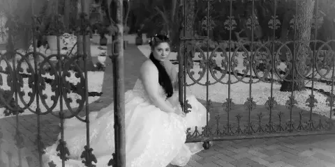 A black and white photograph of a bride in a white wedding dress sitting on a brick path in front of decorative iron gates.