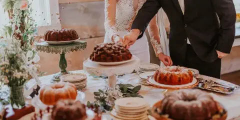A bride and groom cutting a wedding cake at a decorated table with floral arrangements and plates.