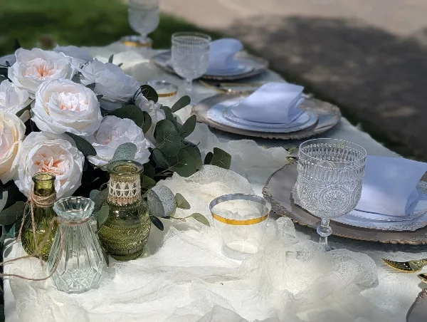 A close-up of a styled event table featuring white roses, greenery, glass vases, and elegant place settings.
