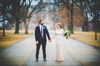 A bride and groom holding hands and smiling while walking down a paved path outdoors.