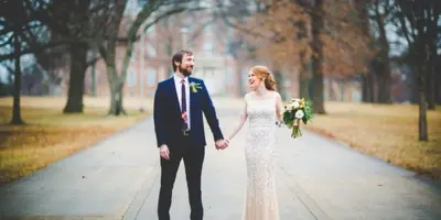 A bride and groom holding hands and smiling while walking down a paved path outdoors.