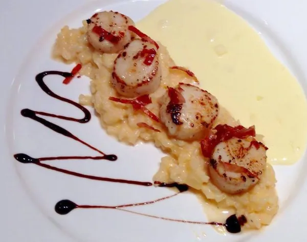 A plated dish featuring seared scallops on a bed of risotto with a yellow sauce and dark balsamic drizzle.