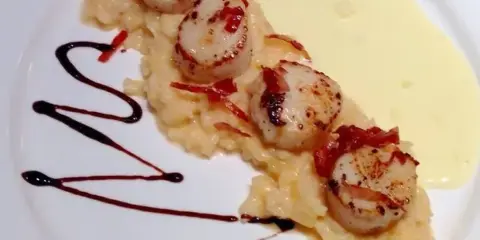 A plated dish featuring seared scallops on a bed of risotto with a yellow sauce and dark balsamic drizzle.