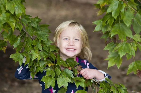 A smiling young girl with blonde hair standing outdoors among green maple leaves.