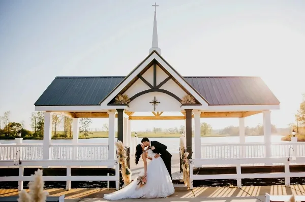 A bride and groom kissing in front of a white outdoor chapel with a steeple, situated on a dock overlooking a lake.