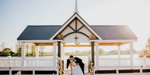A bride and groom kissing in front of a white outdoor chapel with a steeple, situated on a dock overlooking a lake.