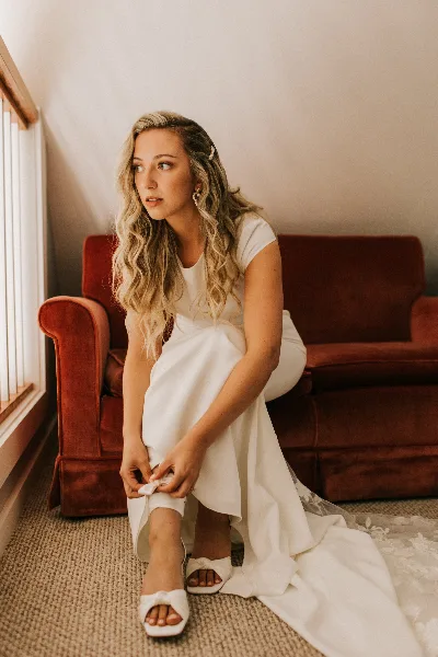 A bride with styled hair and natural makeup sitting on a red couch indoors.