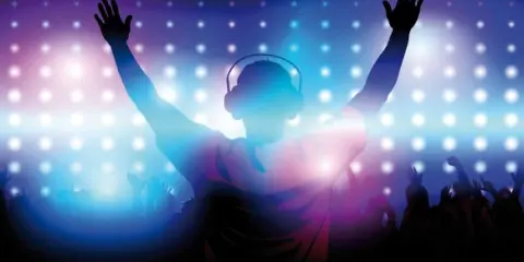 Silhouette of a DJ with raised hands and headphones in front of a crowd and colorful stage lights.