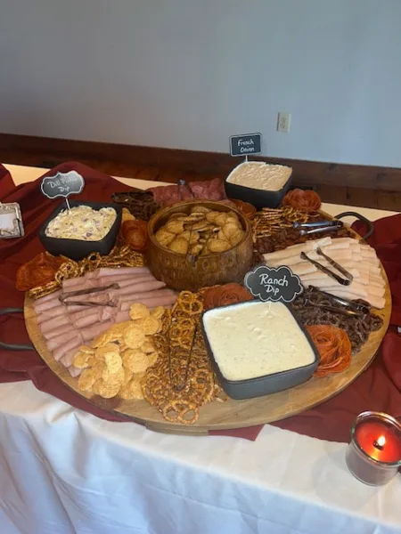 A large wooden board filled with assorted meats, crackers, pretzels, and dips, set on a table with a white tablecloth.