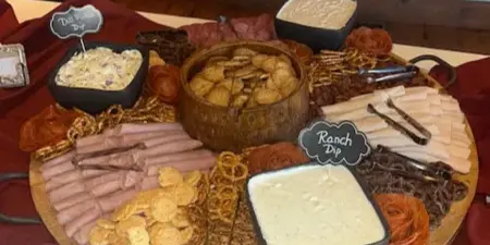 A large wooden board filled with assorted meats, crackers, pretzels, and dips, set on a table with a white tablecloth.