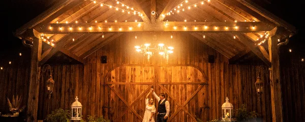 A couple dancing in a rustic wooden barn venue decorated with string lights and a chandelier.