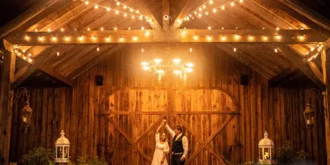 A couple dancing in a rustic wooden barn venue decorated with string lights and a chandelier.