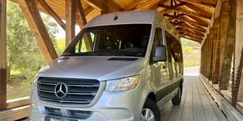 A silver Mercedes-Benz Sprinter van parked under a wooden covered bridge.