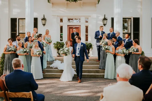 A bride and groom walk down the steps of a white historic building with columns, surrounded by their wedding party.
