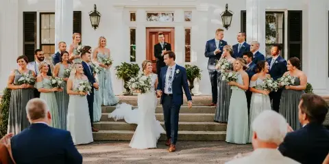 A bride and groom walk down the steps of a white historic building with columns, surrounded by their wedding party.