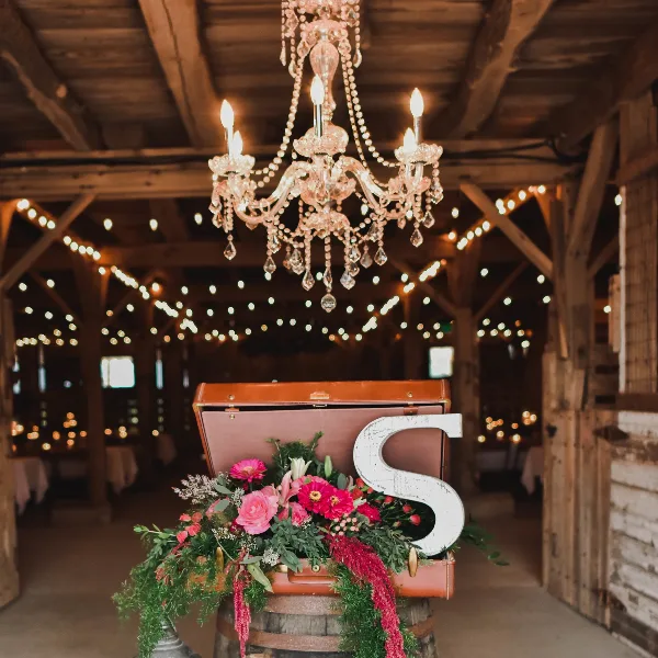 Interior view of a rustic barn wedding venue featuring a crystal chandelier, string lights, and a floral arrangement on a wooden barrel.