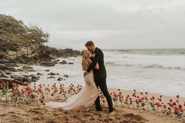 A bride and groom kissing on a beach surrounded by a line of red roses.