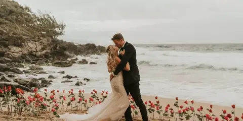 A bride and groom kissing on a beach surrounded by a line of red roses.