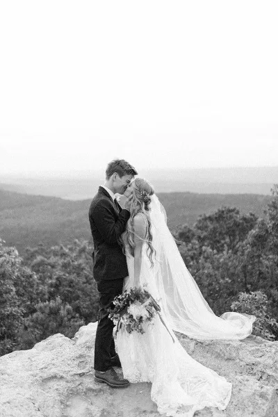 A black and white photograph of a bride and groom kissing on a rocky cliff overlooking a valley.
