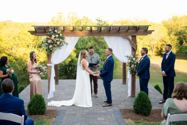 A bride and groom exchanging vows under a wooden pergola with white drapery and floral decorations during an outdoor wedding ceremony.