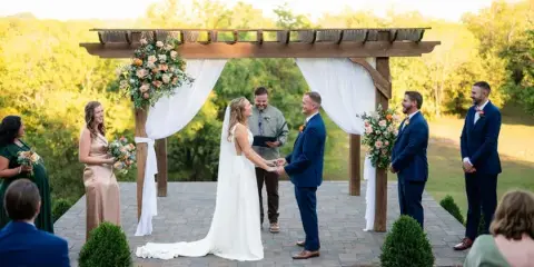 A bride and groom exchanging vows under a wooden pergola with white drapery and floral decorations during an outdoor wedding ceremony.