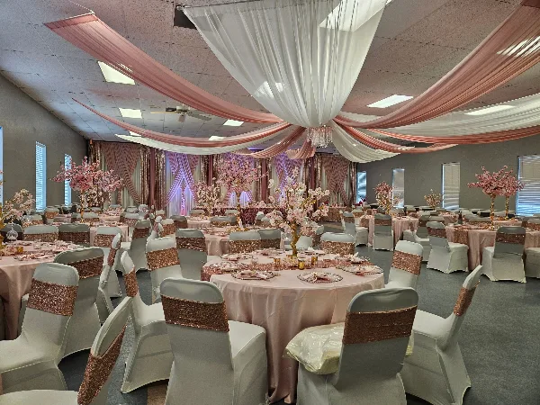 A banquet hall decorated with pink and white ceiling drapes, round tables with pink linens, and chairs with spandex covers and sequined sashes.