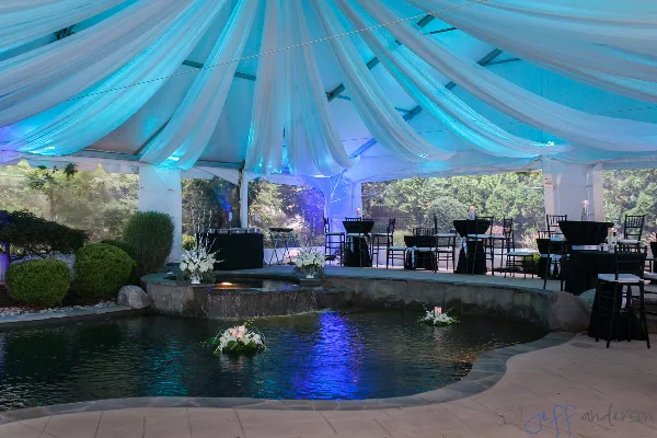 An outdoor event setup featuring a large white tent with blue uplighting, draped ceiling fabric, and tables with black linens and chairs arranged near a water feature.