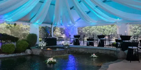 An outdoor event setup featuring a large white tent with blue uplighting, draped ceiling fabric, and tables with black linens and chairs arranged near a water feature.