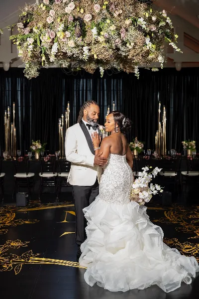 A bride and groom dancing in a ballroom beneath a large floral chandelier installation.