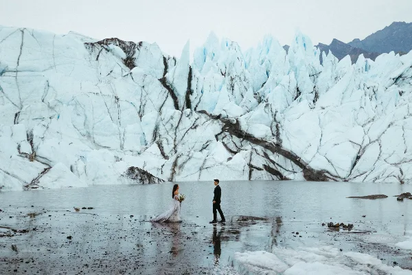 A bride and groom standing in a shallow body of water in front of a massive blue glacier.