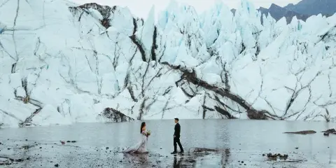 A bride and groom standing in a shallow body of water in front of a massive blue glacier.